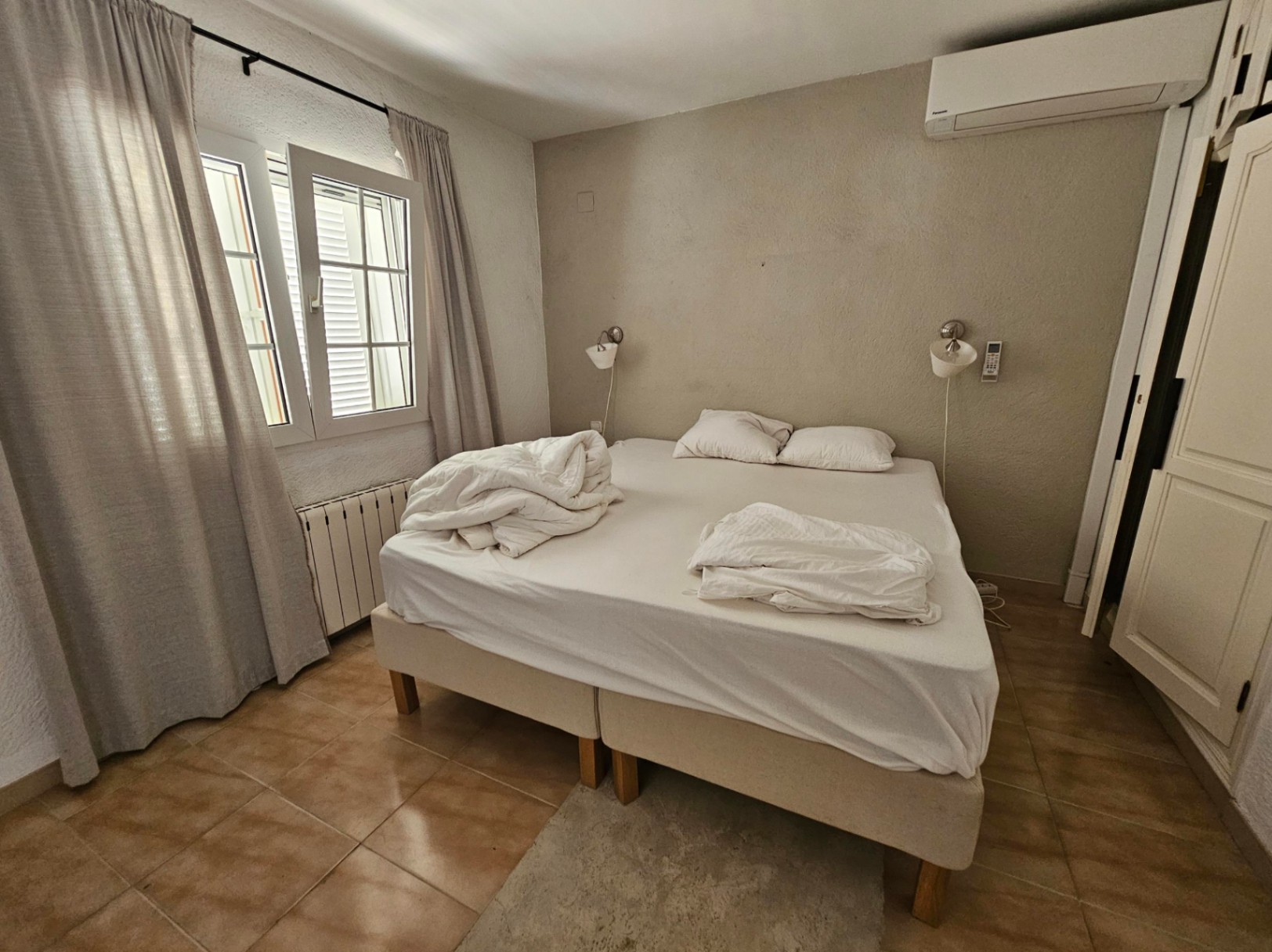 Dormitorio con cama doble, cortinas de lino y suelo de baldosas rústicas.
