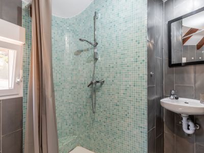 Detalle de baño con ducha de mosaico turquesa, lavabo suspendido y azulejos grises.