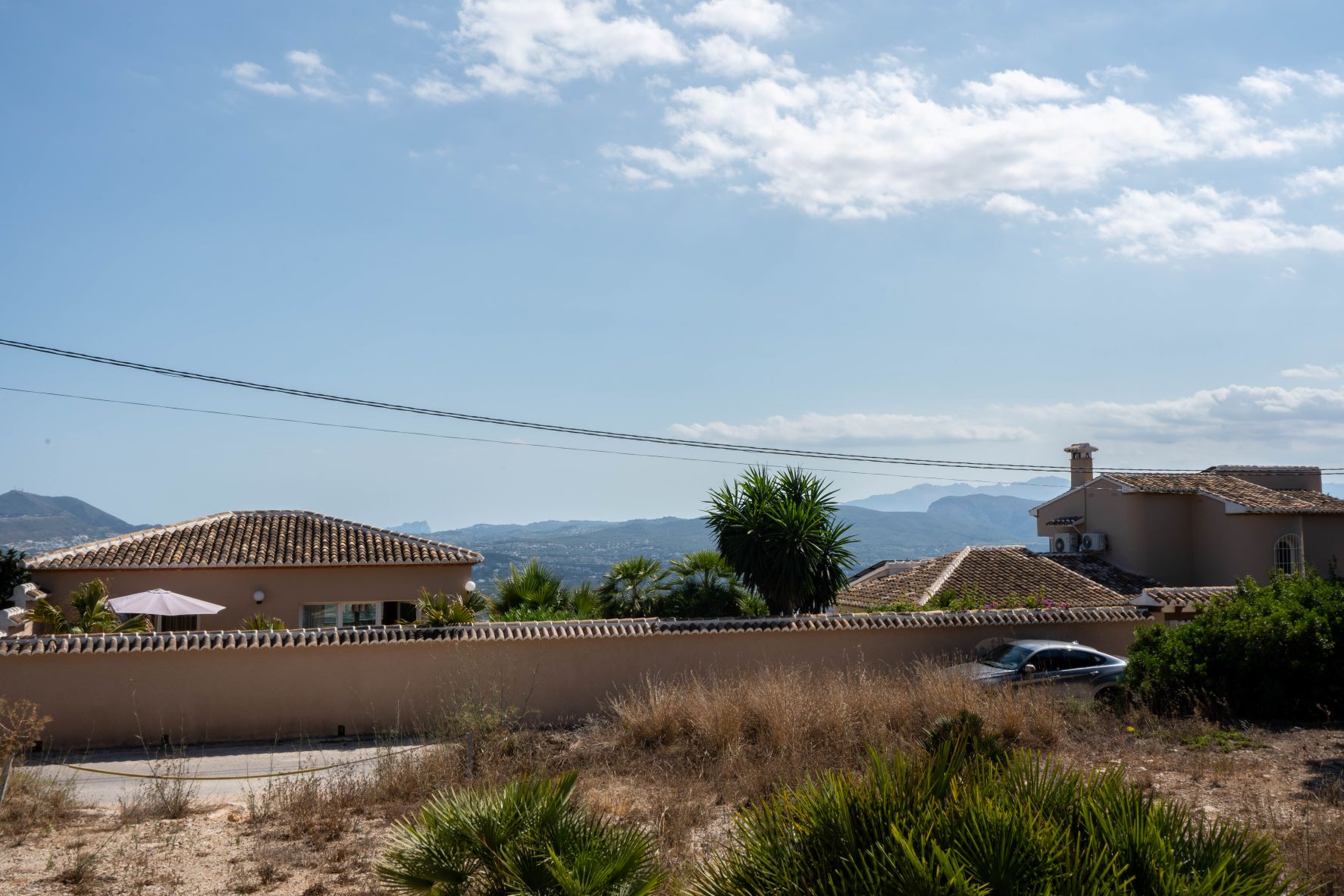 Vista de la parcela con casas vecinas, vegetación y montañas al fondo bajo un cielo parcialmente nublado.