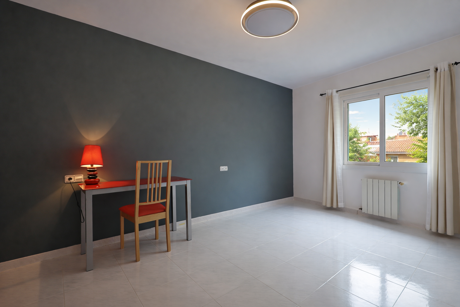 Habitación luminosa con escritorio rojo y pared gris plomo, baldosas color crema y luz de techo circular.