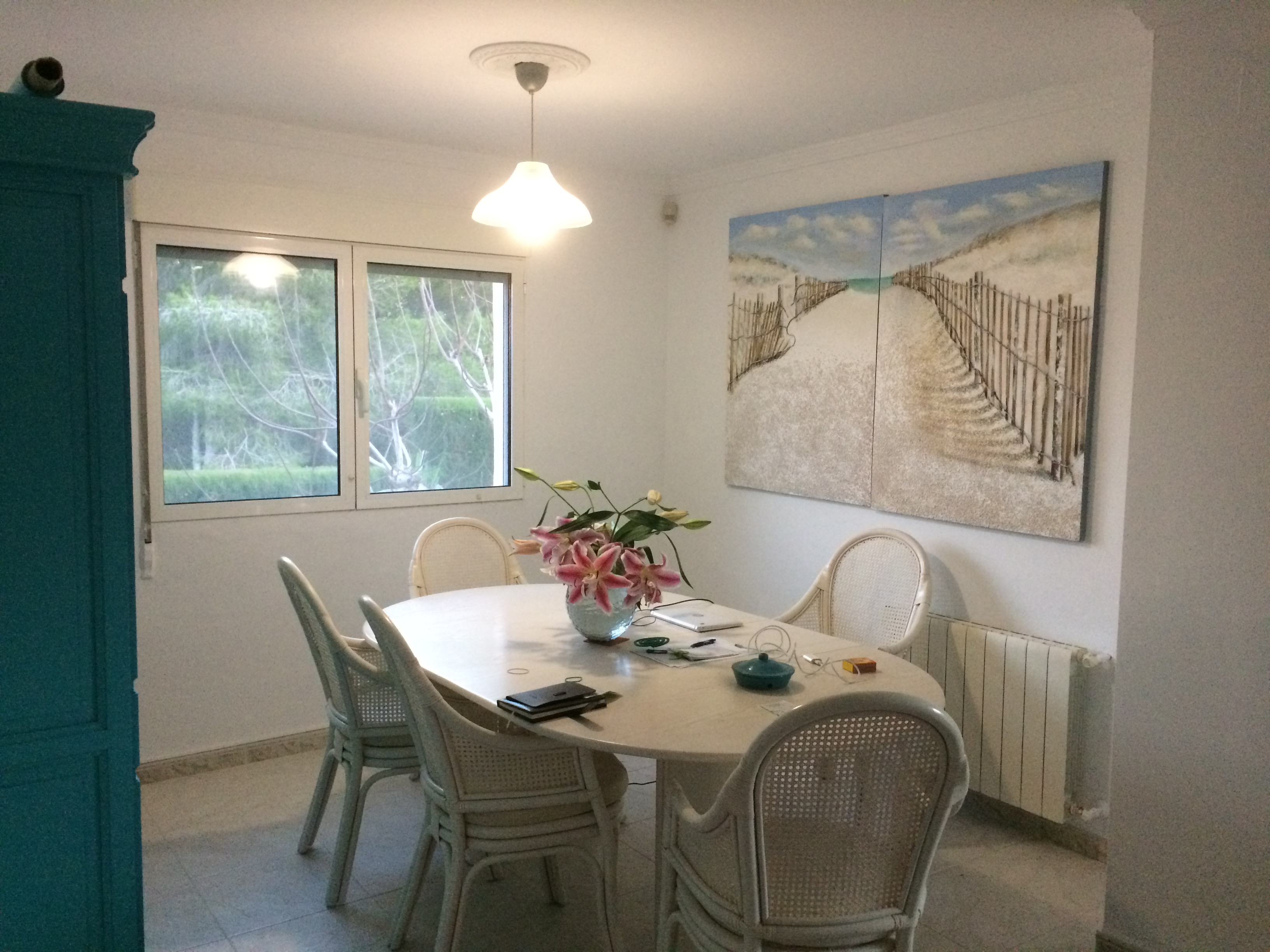 Comedor luminoso con mesa ovalada de piedra y sillas de mimbre, decorado con un cuadro de playa.