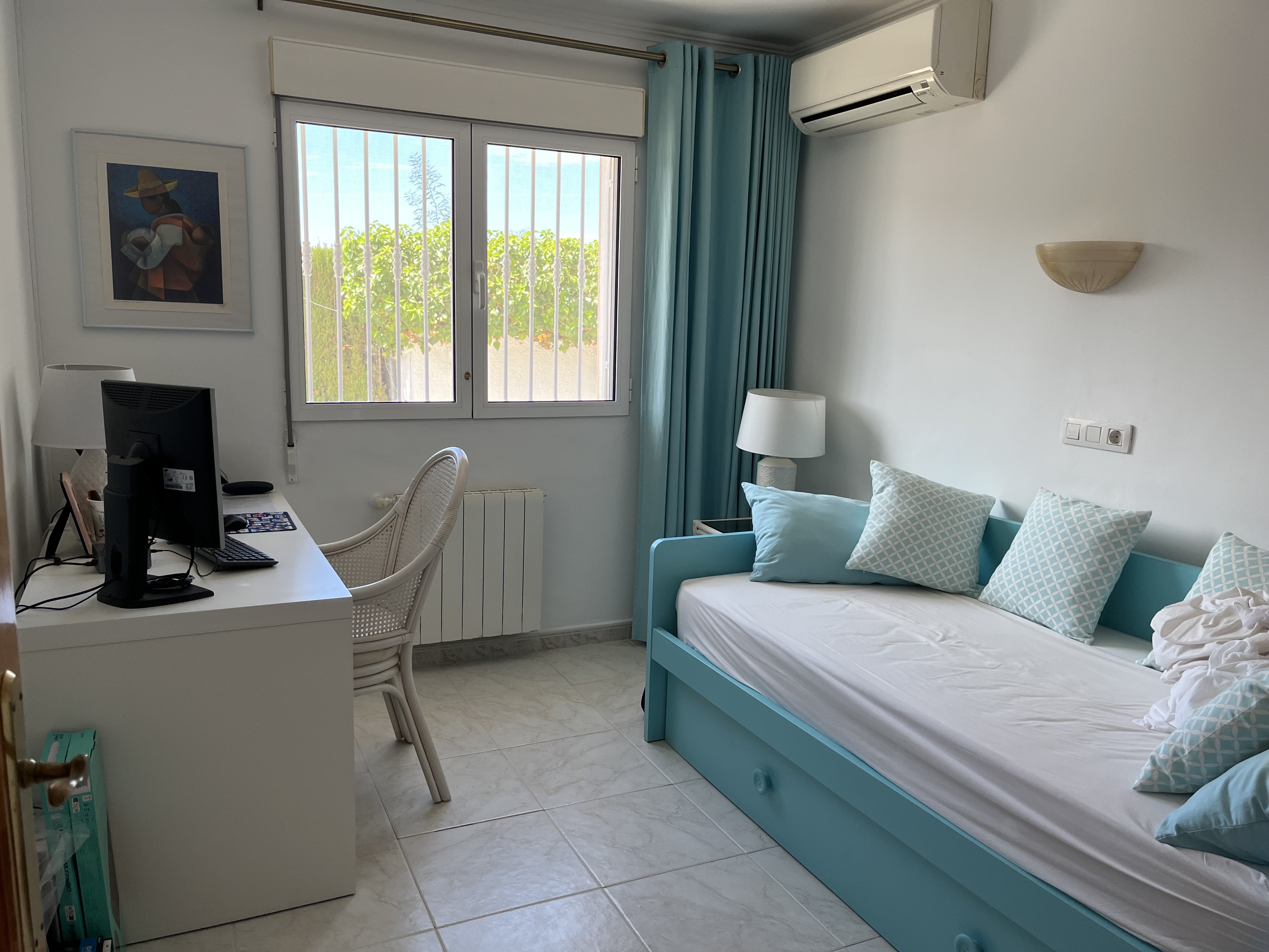Habitación luminosa con cama turquesa, escritorio blanco y vistas al jardín.