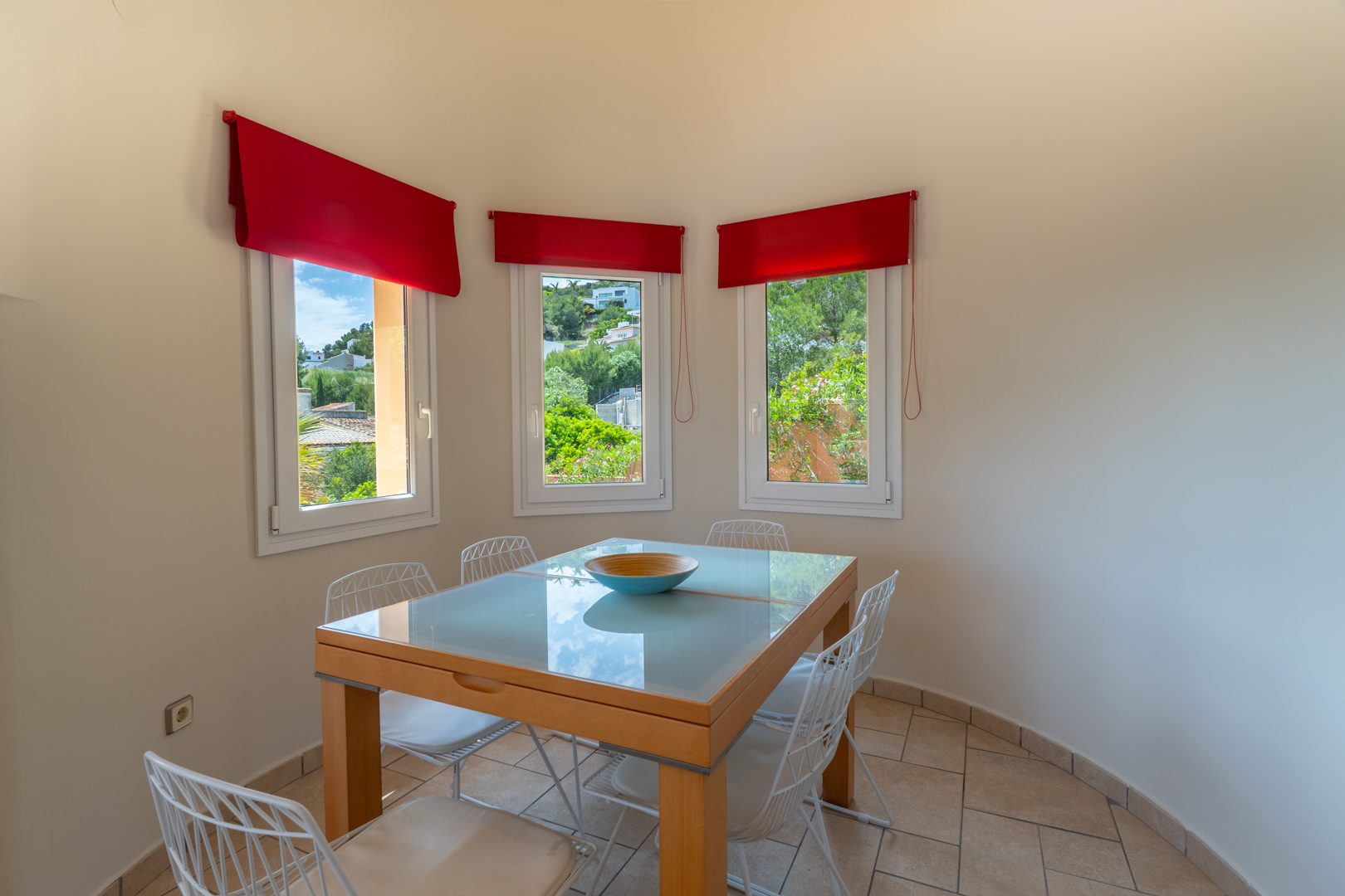 Comedor luminoso con mesa de cristal y vistas al exterior a través de ventanas con cortinas rojas.