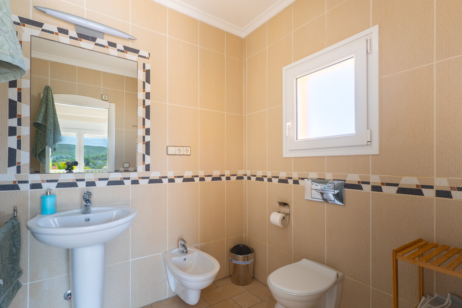 Detalle de baño con azulejos color crema y cenefa decorativa. Incluye lavabo con pedestal, bidé e inodoro.