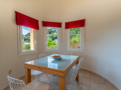 Comedor luminoso con mesa de cristal y vistas al exterior a través de ventanas con cortinas rojas.