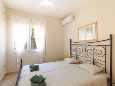 Dormitorio luminoso con camas individuales, cabecero de hierro forjado y vistas al exterior a través de cortinas blancas.
