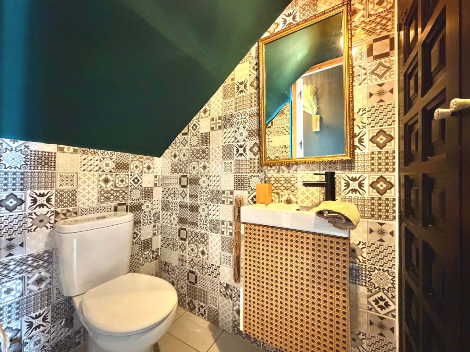 Aseo con azulejos estampados, techo verde oscuro y lavabo con frontal de mimbre.