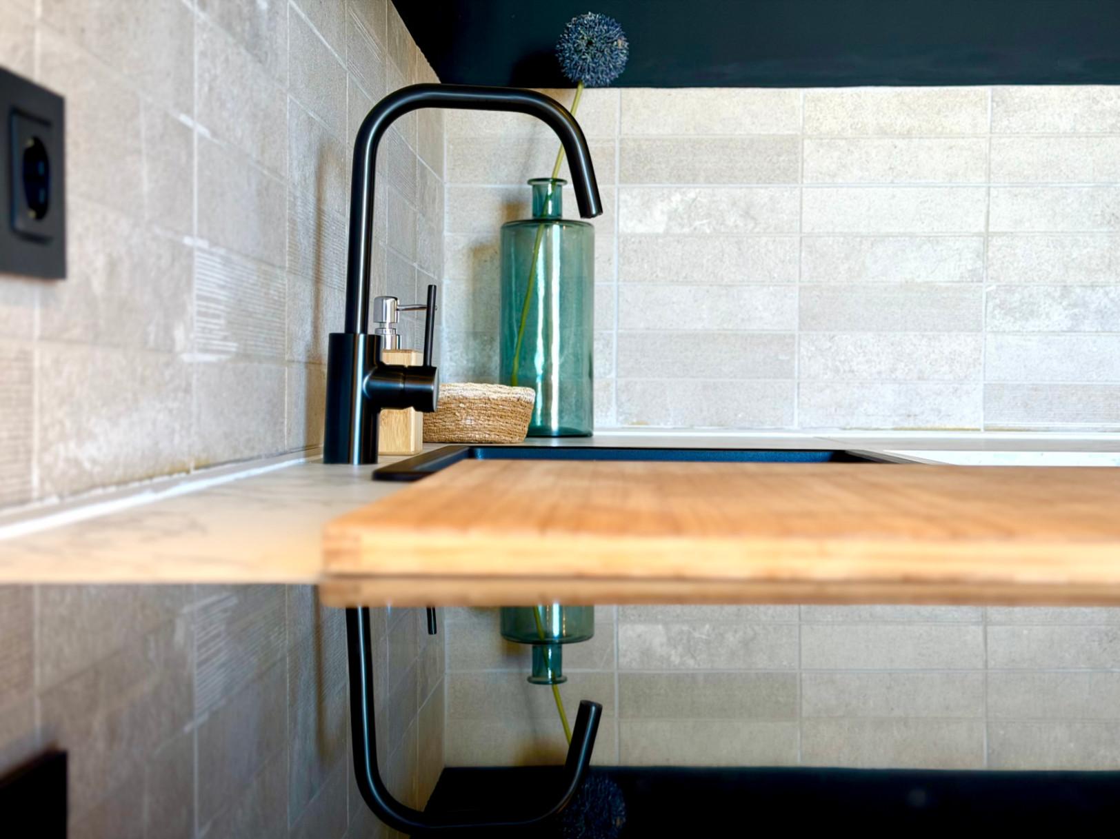 Detalle de la cocina con grifo negro, tabla de cortar de madera y azulejos color crema.