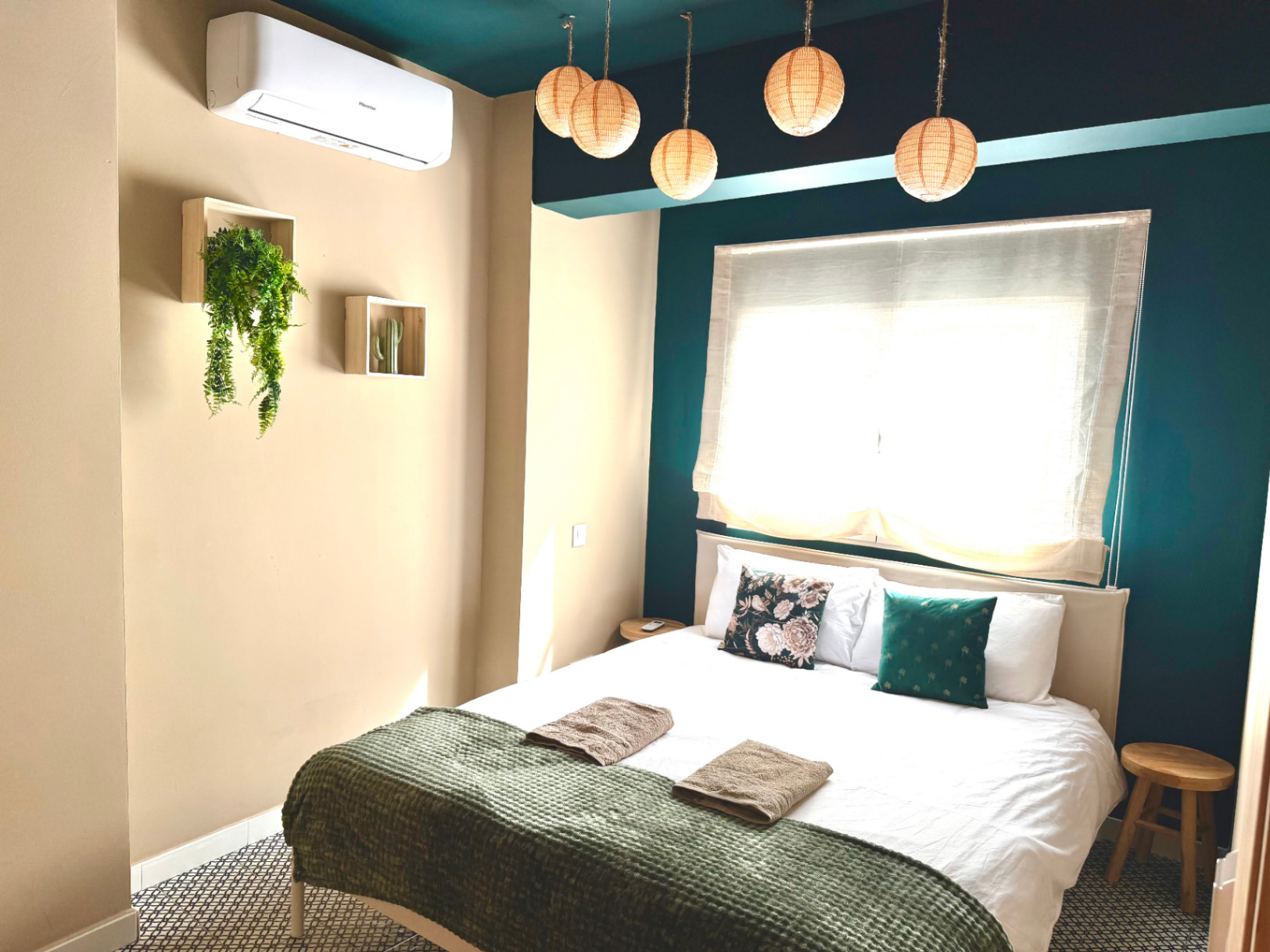 Dormitorio luminoso con paredes beige, acento turquesa y lámparas colgantes de mimbre. Cama doble con manta verde oliva.