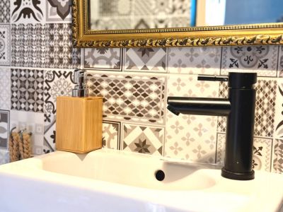 Detalle de lavabo con grifo negro mate y dispensador de jabón de bambú. Azulejos decorativos en pared y espejo con marco dorado.