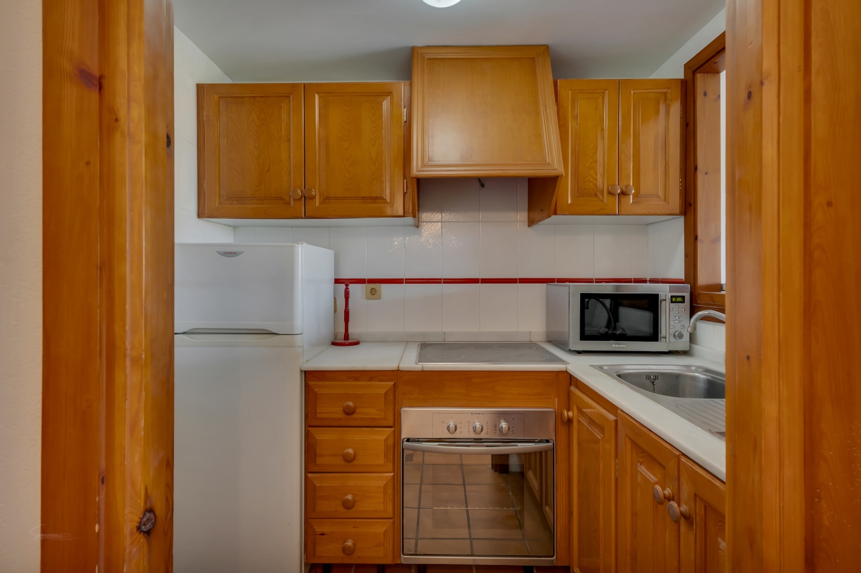 Cocina funcional con muebles de madera, electrodomésticos básicos y azulejos blancos con detalles rojos.