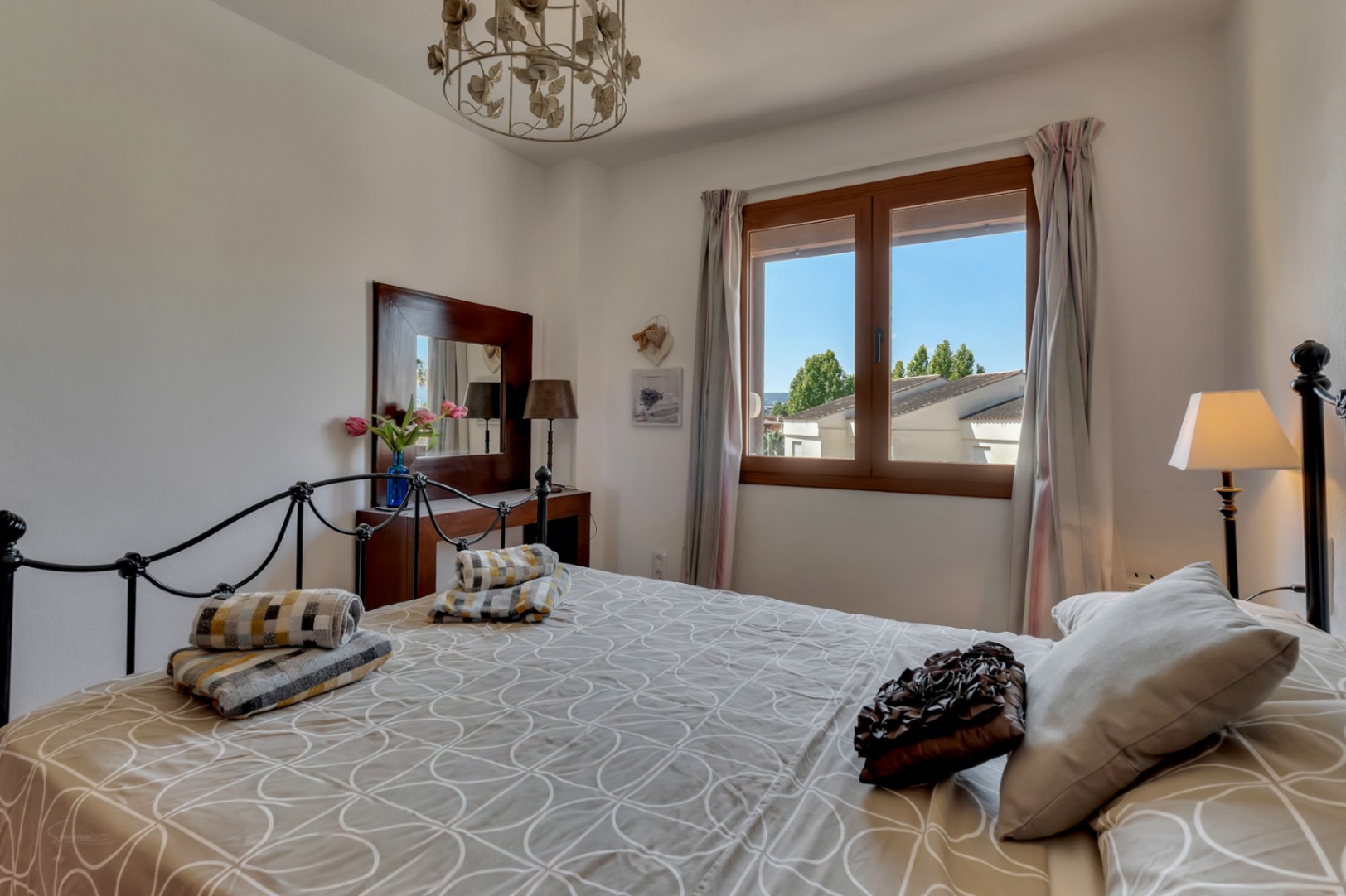 Dormitorio luminoso con cama de hierro forjado, ropa de cama estampada y ventana de madera con vistas al exterior.