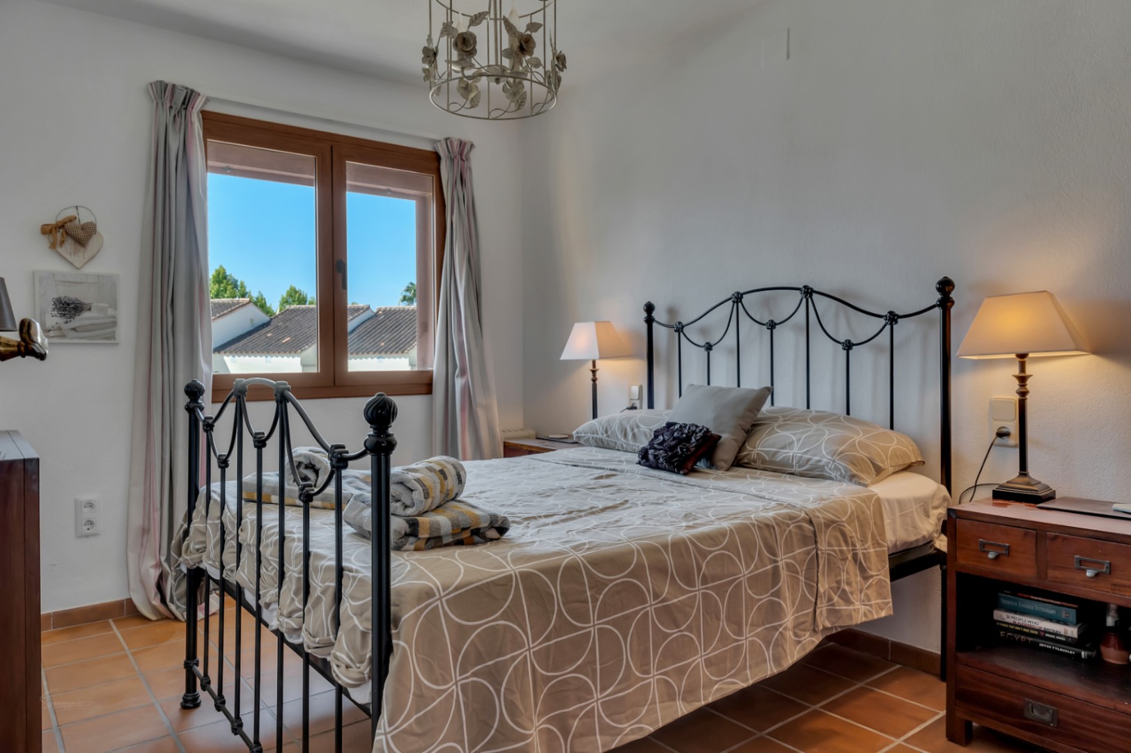 Dormitorio luminoso con cama de hierro forjado, ventana con vistas y suelo de terracota.