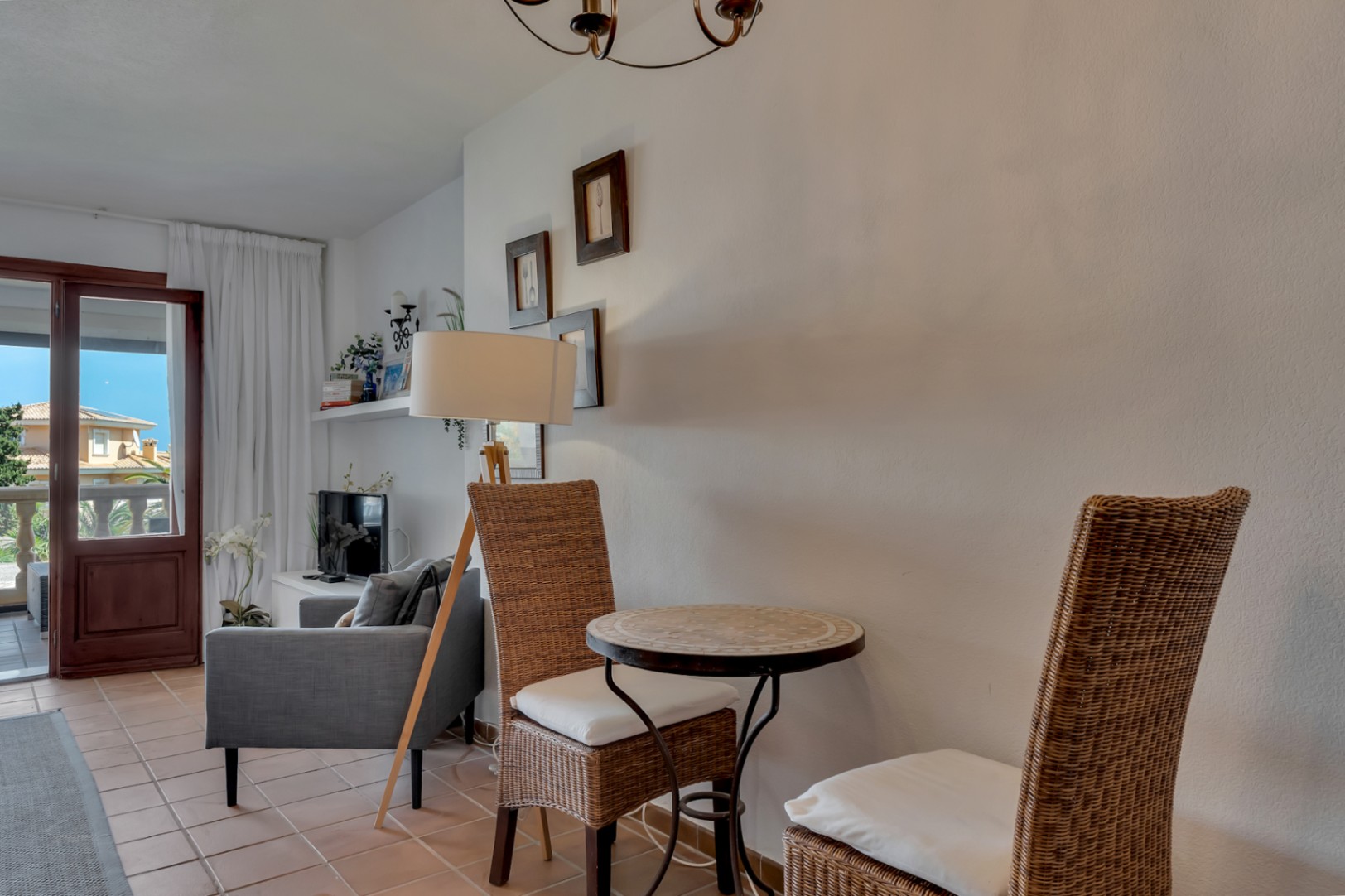 Rincón acogedor con mesa de mosaico y sillas de mimbre, iluminado por una lámpara de pie y luz natural desde la terraza.