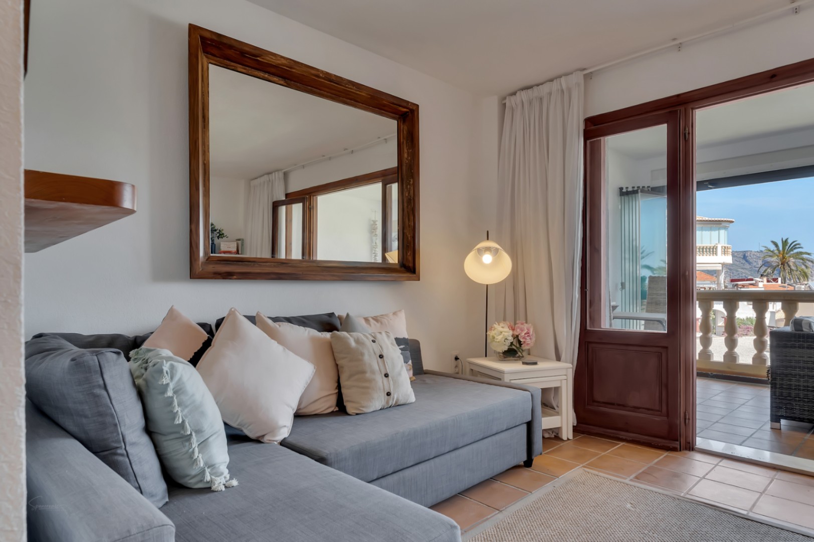 Salón con sofá gris, espejo de madera y acceso a terraza con vistas al mar en Jávea.