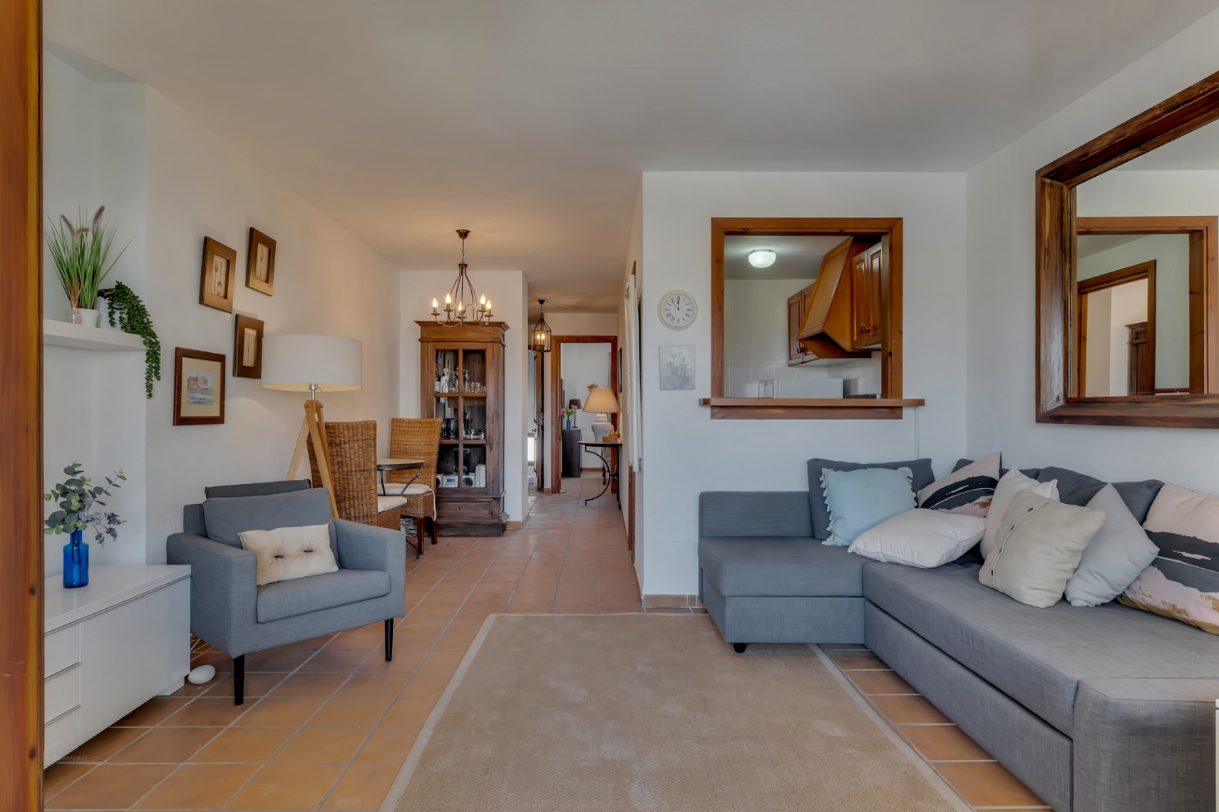 Salón con sofá gris, espejo de madera y vistas a la cocina a través de una ventana interior.