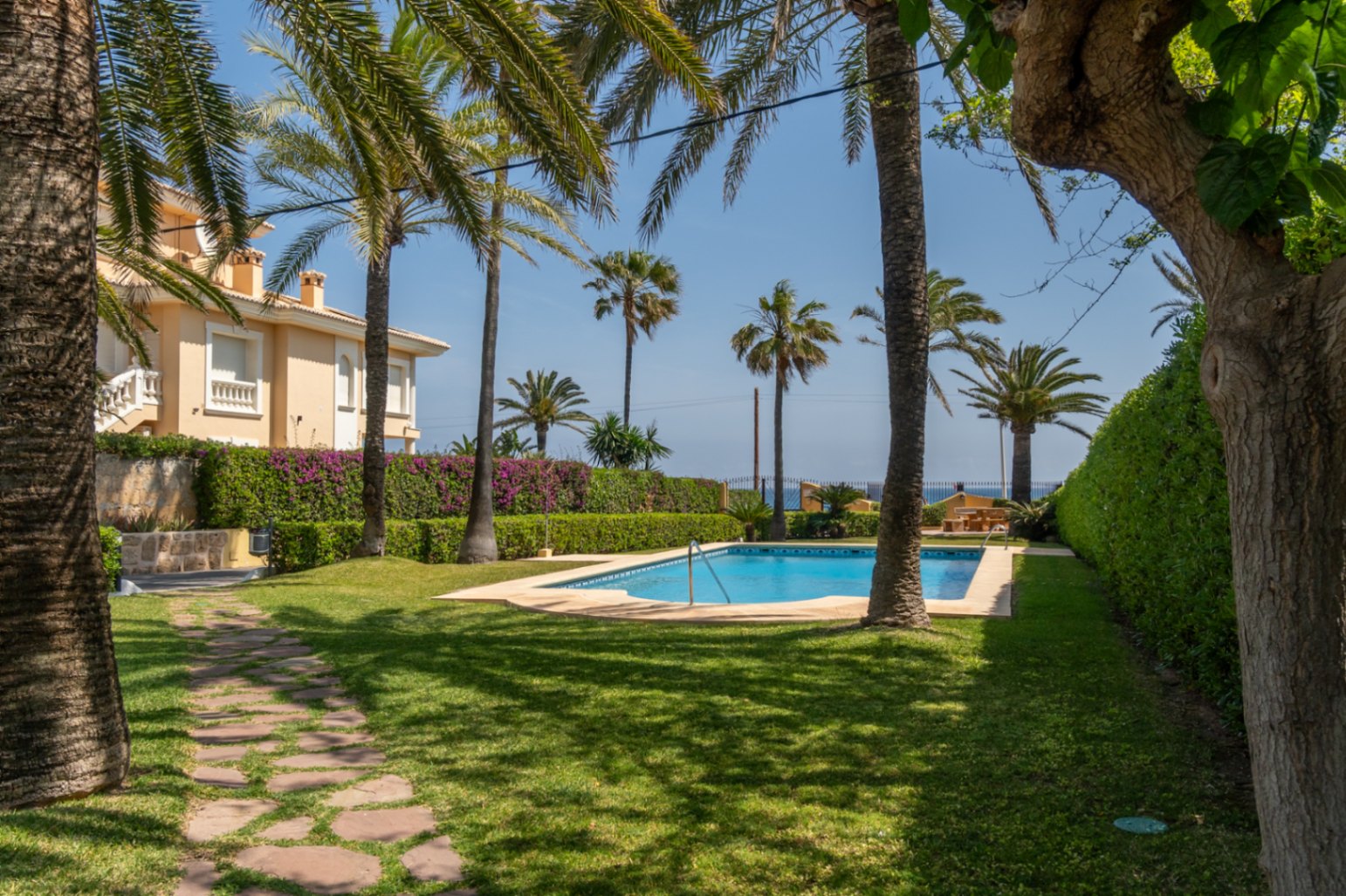 Vista de la piscina comunitaria rodeada de césped y palmeras en una urbanización en Jávea.