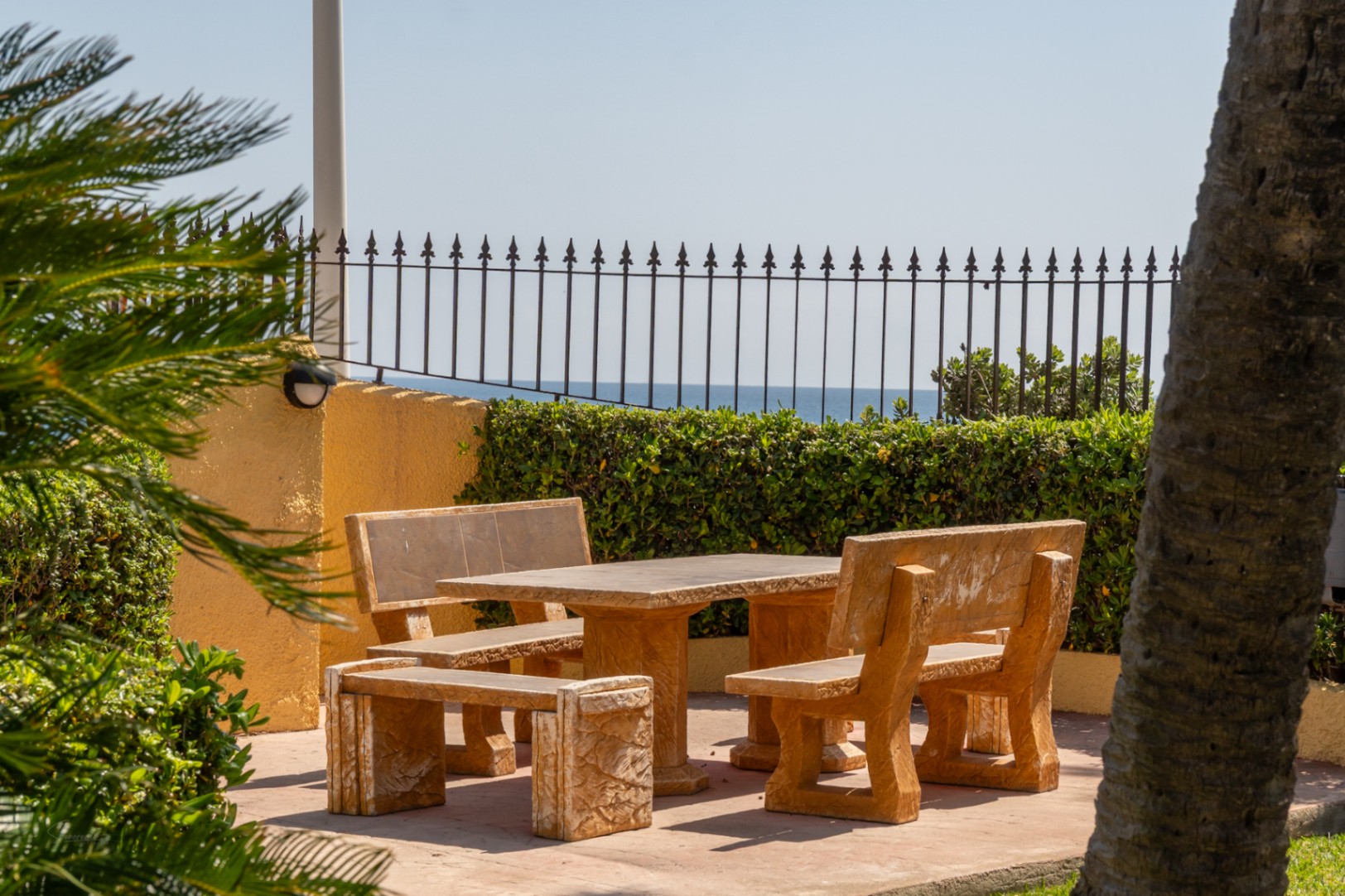 Zona de estar al aire libre con mesa y bancos de piedra, jardines mediterráneos y vistas al mar.