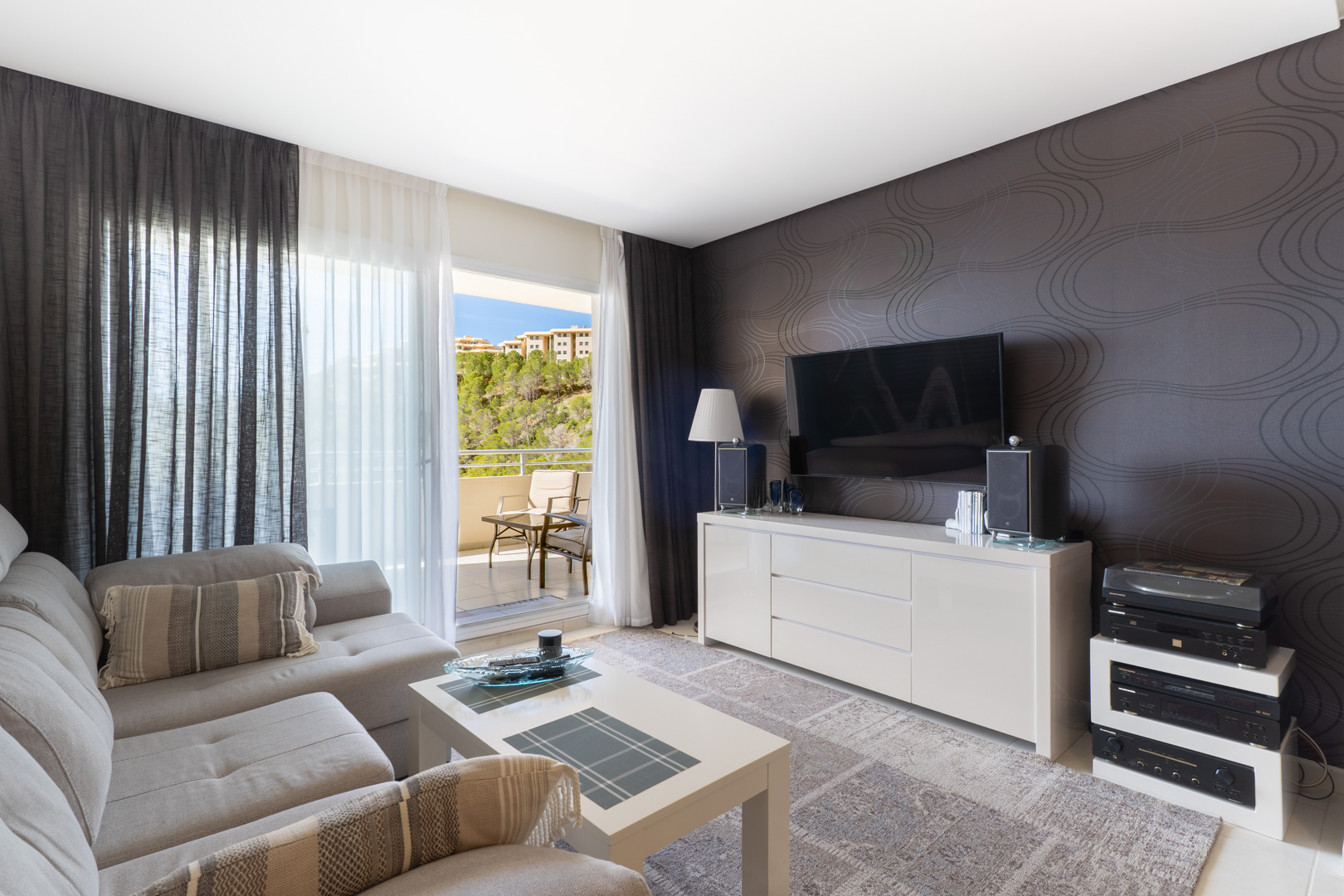 Amplio salón con sofá seccional gris claro, mesa de centro blanca y mueble de TV frente a una pared decorada. Puertas correderas acceden a un balcón con vistas a la colinas. Amplio salón con sofá seccional gris claro, mesa de centro blanca y mueble de TV frente a una pared decorada. Puertas correderas acceden a un balcón con vistas a la colinas.