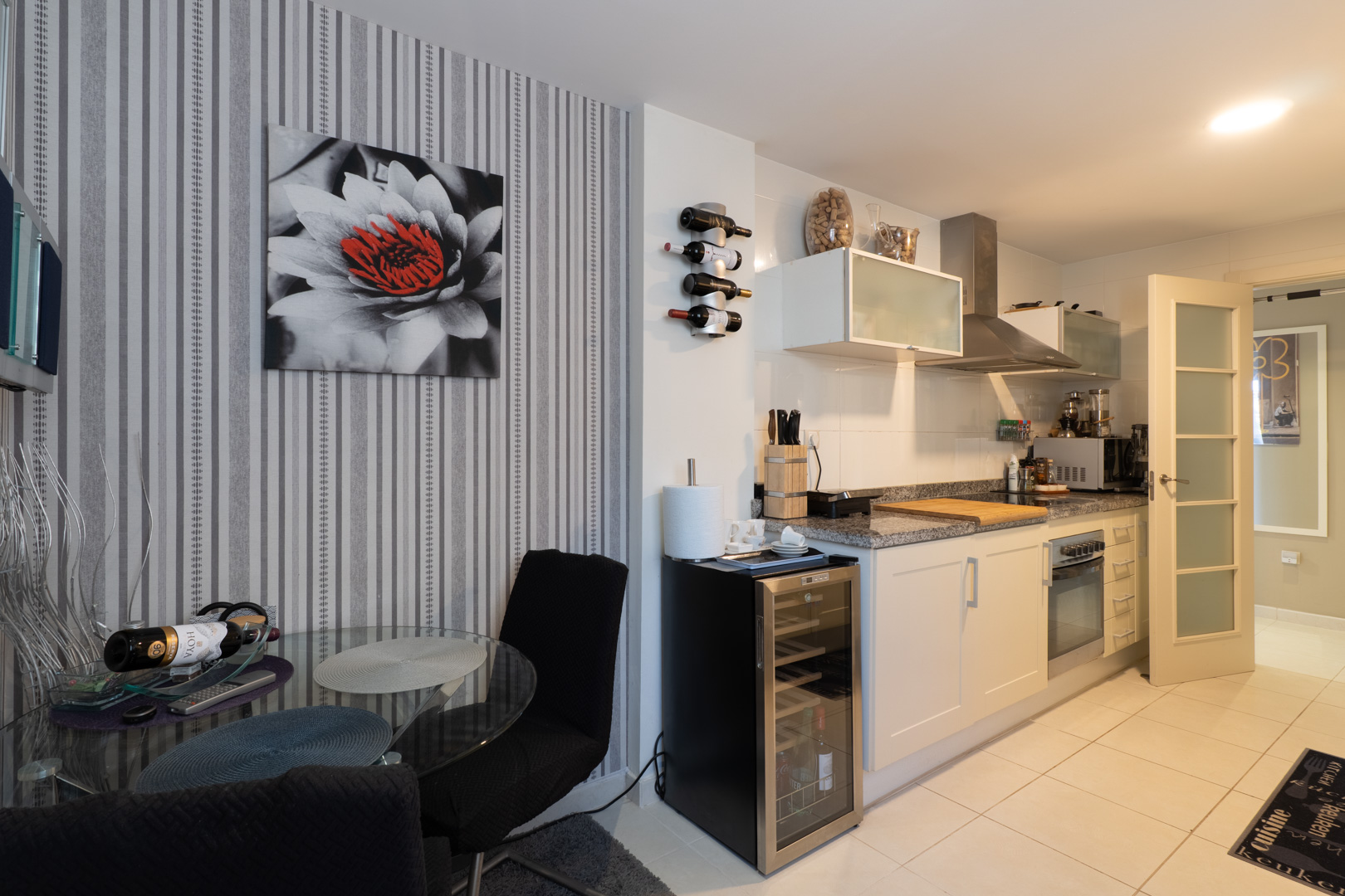 Cocina moderna con encimera de granito, electrodomésticos integrados y botellero. Incluye una vinoteca y mesa de comedor de cristal junto a una pared con papel pintado a rayas. Cocina moderna con encimera de granito, electrodomésticos integrados y botellero. Incluye una vinoteca y mesa de comedor de cristal junto a una pared con papel pintado a rayas.