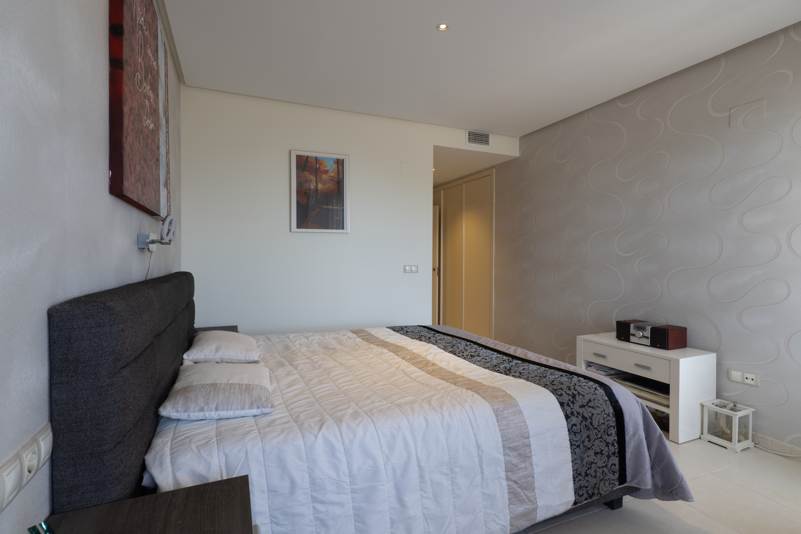 Dormitorio principal con cama doble, cabecero tapizado gris y edredón blanco. Destaca una pared con papel pintado de ondas y armarios empotrados visibles al fondo. Dormitorio principal con cama doble, cabecero tapizado gris y edredón blanco. Destaca una pared con papel pintado de ondas y armarios empotrados visibles al fondo.