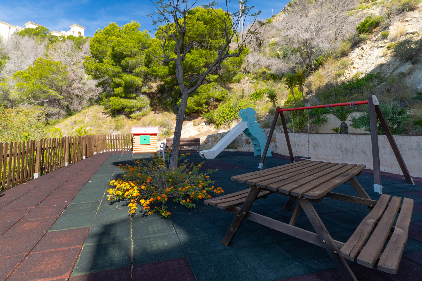 Parque infantil con mesa de picnic de madera, columpios y tobogán sobre suelo de caucho multicolor. Una casita de juegos y valla de madera se aprecian, con una ladera verde al fondo. Parque infantil con mesa de picnic de madera, columpios y tobogán sobre suelo de caucho multicolor. Una casita de juegos y valla de madera se aprecian, con una ladera verde al fondo.