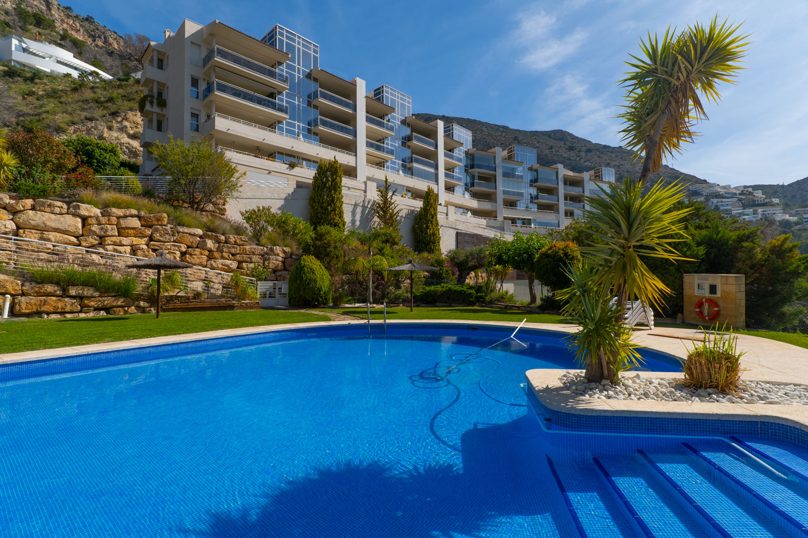 Piscina comunitaria de mosaico azul con acceso escalonado, rodeada de césped y vegetación mediterránea, frente a un edificio moderno con balcones de cristal en la ladera. Piscina comunitaria de mosaico azul con acceso escalonado, rodeada de césped y vegetación mediterránea, frente a un edificio moderno con balcones de cristal en la ladera.