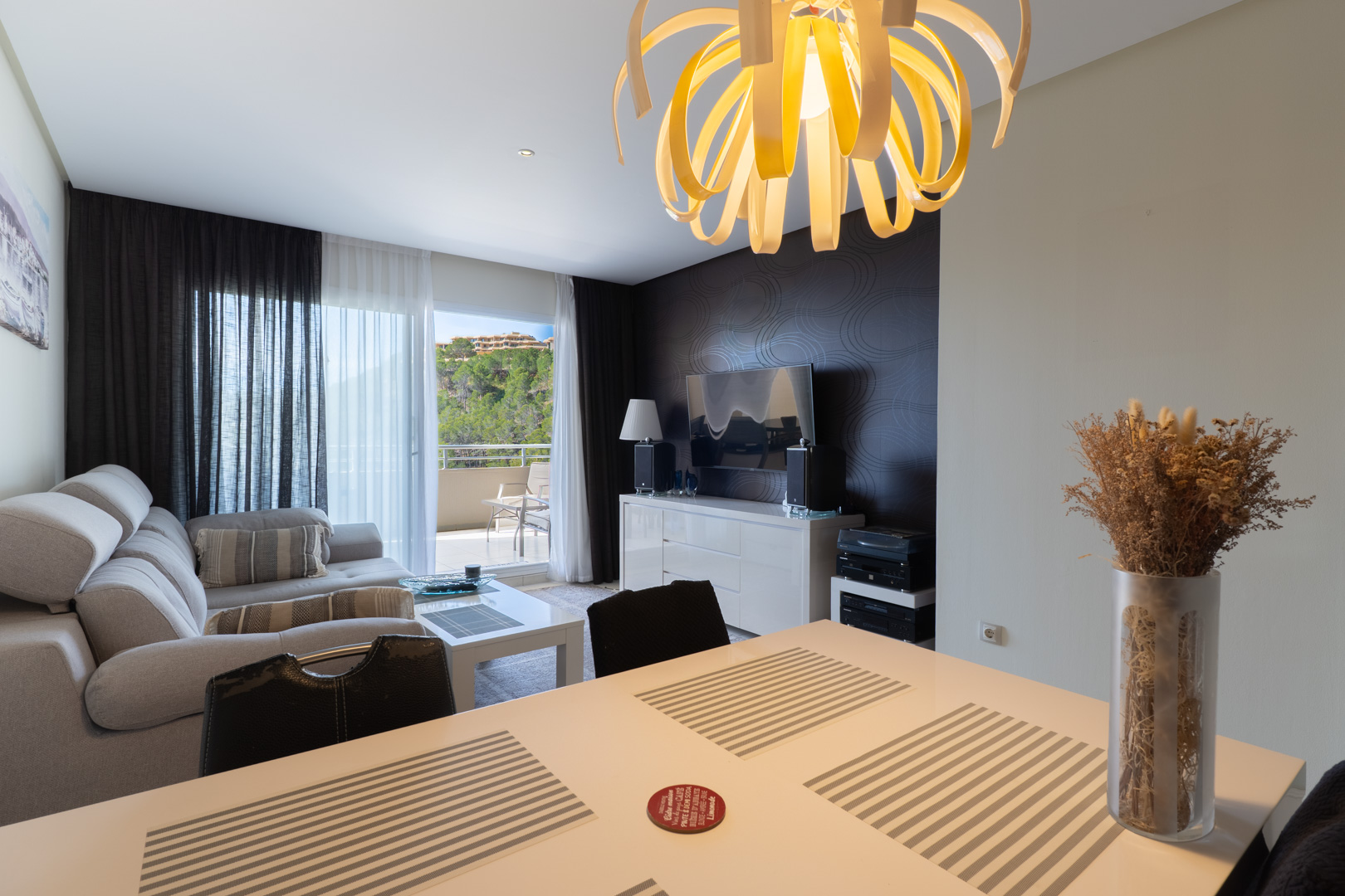 Salón-comedor de planta abierta con un distintivo candelabro en espiral, sofá seccional gris claro y mesa de comedor blanca. Puerta acristalada al balcón con vistas a la vegetación. Salón-comedor de planta abierta con un distintivo candelabro en espiral, sofá seccional gris claro y mesa de comedor blanca. Puerta acristalada al balcón con vistas a la vegetación.