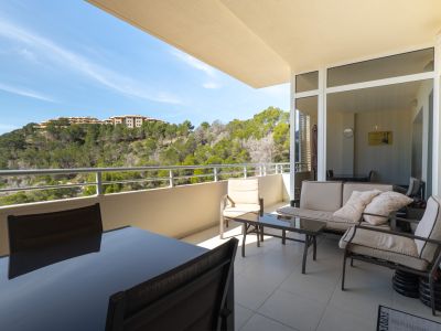 Amplia terraza exterior con mobiliario de comedor y salón, ofreciendo vistas a una ladera verde con pinos y edificios lejanos.