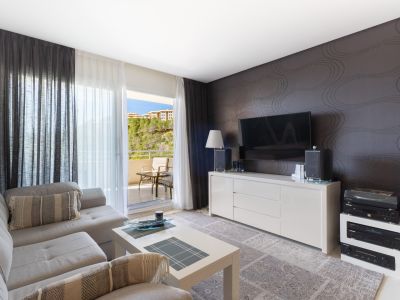 Amplio salón con sofá seccional gris claro, mesa de centro blanca y mueble de TV frente a una pared decorada. Puertas correderas acceden a un balcón con vistas a la colinas.