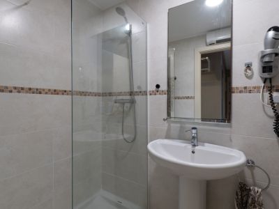 Baño con ducha acristalada, azulejos claros y franja decorativa de mosaico. Incluye lavabo de pedestal, espejo y secador de pelo mural.