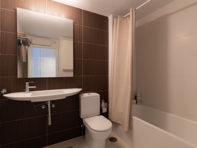 Baño funcional con lavabo suspendido, inodoro y bañera con cortina. Combina azulejos rectangulares marrones oscuros con baldosas claras en las paredes.