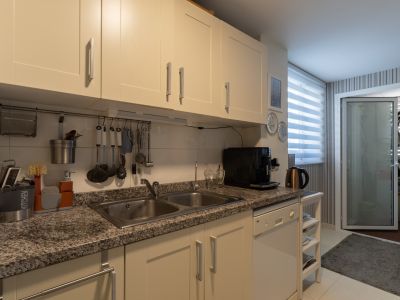 Cocina funcional con armarios color crema, encimera de granito moteado con fregadero doble de acero inoxidable y lavavajillas integrado blanco. Luz natural a través de la ventana con persianas.