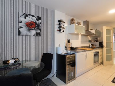 Cocina moderna con encimera de granito, electrodomésticos integrados y botellero. Incluye una vinoteca y mesa de comedor de cristal junto a una pared con papel pintado a rayas.