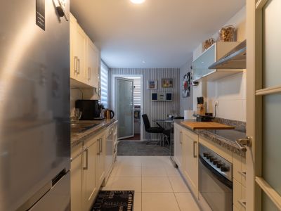 Cocina tipo galera equipada con muebles claros, encimeras de granito y electrodomésticos de acero inoxidable, incluyendo nevera y horno. Al fondo, un pequeño rincón comedor con mesa de cristal.