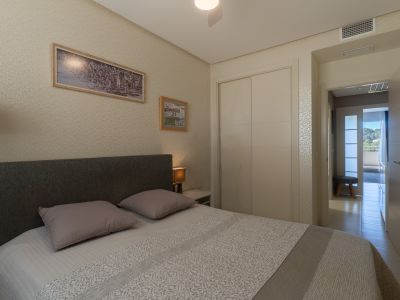 Dormitorio con cama doble, cabecero de tela, armario empotrado y papel pintado. Se observa ventilación de aire acondicionado y un pasillo luminoso hacia el exterior.
