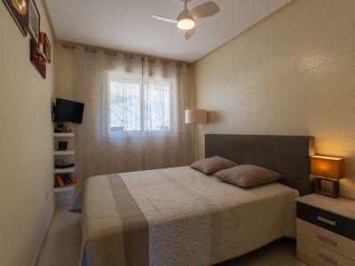 Dormitorio con cama doble, cabecero tapizado y mesita de noche con lámpara. La estancia cuenta con ventana con cortinas, ventilador de techo y TV mural.