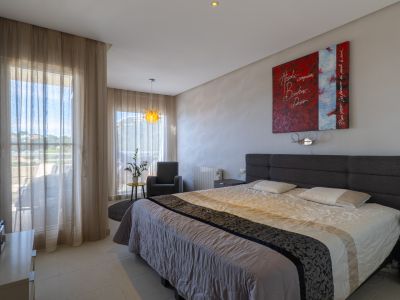 Dormitorio luminoso con cama doble, cabecero tapizado gris y acceso directo a una terraza. El suelo de baldosas claras y una lámpara colgante decorativa complementan la luz natural.
