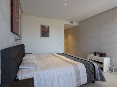 Dormitorio principal con cama doble, cabecero tapizado gris y edredón blanco. Destaca una pared con papel pintado de ondas y armarios empotrados visibles al fondo.