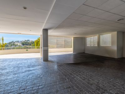 Espaciosa área cubierta con suelo de adoquines oscuros y techo mixto. Ofrece una vista abierta a las colinas verdes y edificios distantes, con luz natural y ventanas con lamas.