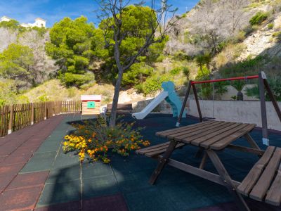 Parque infantil con mesa de picnic de madera, columpios y tobogán sobre suelo de caucho multicolor. Una casita de juegos y valla de madera se aprecian, con una ladera verde al fondo.