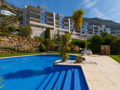 Piscina comunitaria de azulejos azules con escalones, rodeada por un solárium pavimentado y césped, con el edificio de apartamentos moderno en la ladera.