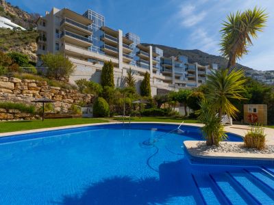 Piscina comunitaria de mosaico azul con acceso escalonado, rodeada de césped y vegetación mediterránea, frente a un edificio moderno con balcones de cristal en la ladera.