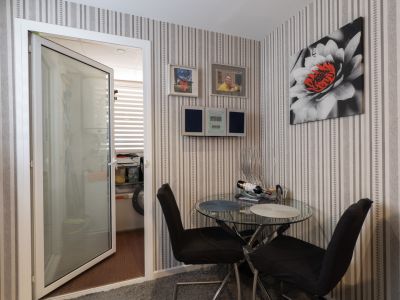 Rincón acogedor con mesa de cristal y sillas negras, adornado con papel pintado a rayas y un cuadro floral. Una puerta abierta revela un área de lavandería con luz natural.