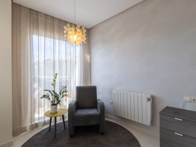 Rincón luminoso con sillón gris, mesa auxiliar amarilla con planta, y lámpara colgante decorativa. Ventanal con cortinas claras y radiador eléctrico.