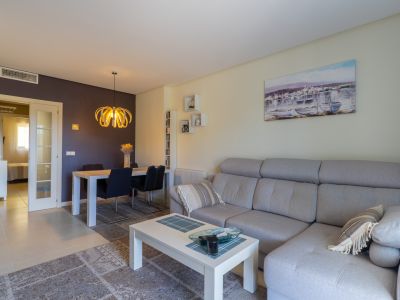 Sala de estar-comedor con sofá seccional gris, mesa de centro blanca y alfombra. Destaca la pared de acento oscura y la lámpara colgante moderna sobre el comedor. Puerta acristalada visible.