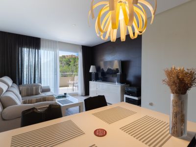 Salón-comedor de planta abierta con un distintivo candelabro en espiral, sofá seccional gris claro y mesa de comedor blanca. Puerta acristalada al balcón con vistas a la vegetación.