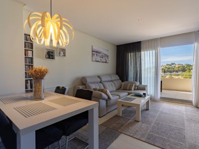 Salón-comedor luminoso con mesa cuadrada, sofá gris y lámpara de diseño. Acceso a terraza con vistas a las colinas y estantería integrada.