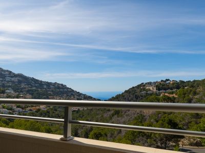 Vista panorámica desde la terraza con barandilla metálica, mostrando colinas verdes con casas y el mar Mediterráneo al fondo bajo cielo azul.