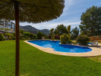 Zona de piscina comunitaria con sombrilla de paja, césped verde y vegetación mediterránea, con colinas y un edificio de apartamentos al fondo.